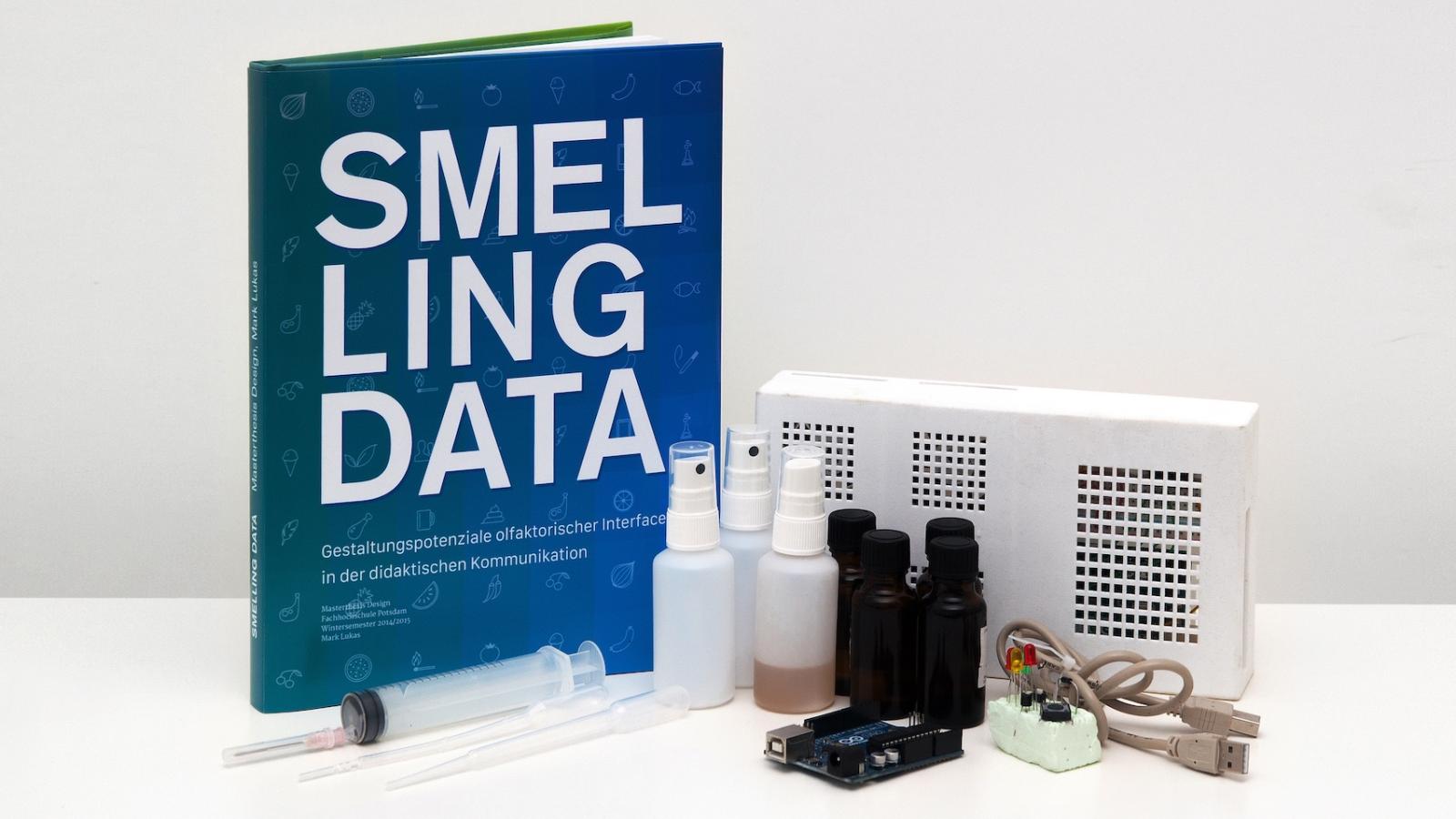 Aufgestelltes Buch mit der Aufschrift "Smelling Data". Daneben verschiedene Gegenst?nde wie kleine Sprhflaschen und USB-Kabel. 