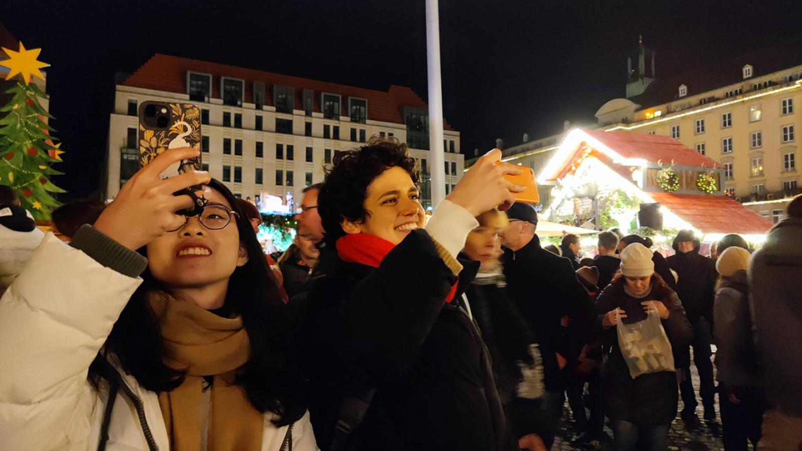 Zwei Personen stehen in der unteren linken Ecke des Bildes und halten ihre Handys in die H?he als wrden sie fotografieren. Sie stehen auf einem Weihnachtsmarkt. Der Himmel ist dunkel aber die Lichter vom Markt gl?nzen hell.