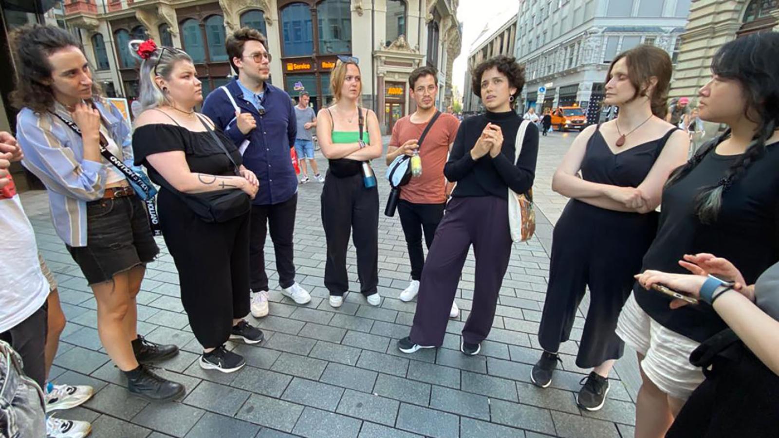 Ungef?hr zehn Menschen stehen in einem Halbkreis auf einem Platz in Leipzig und schauen auf eine Person die Teil des Kreises ist und etwas erz?hlt.