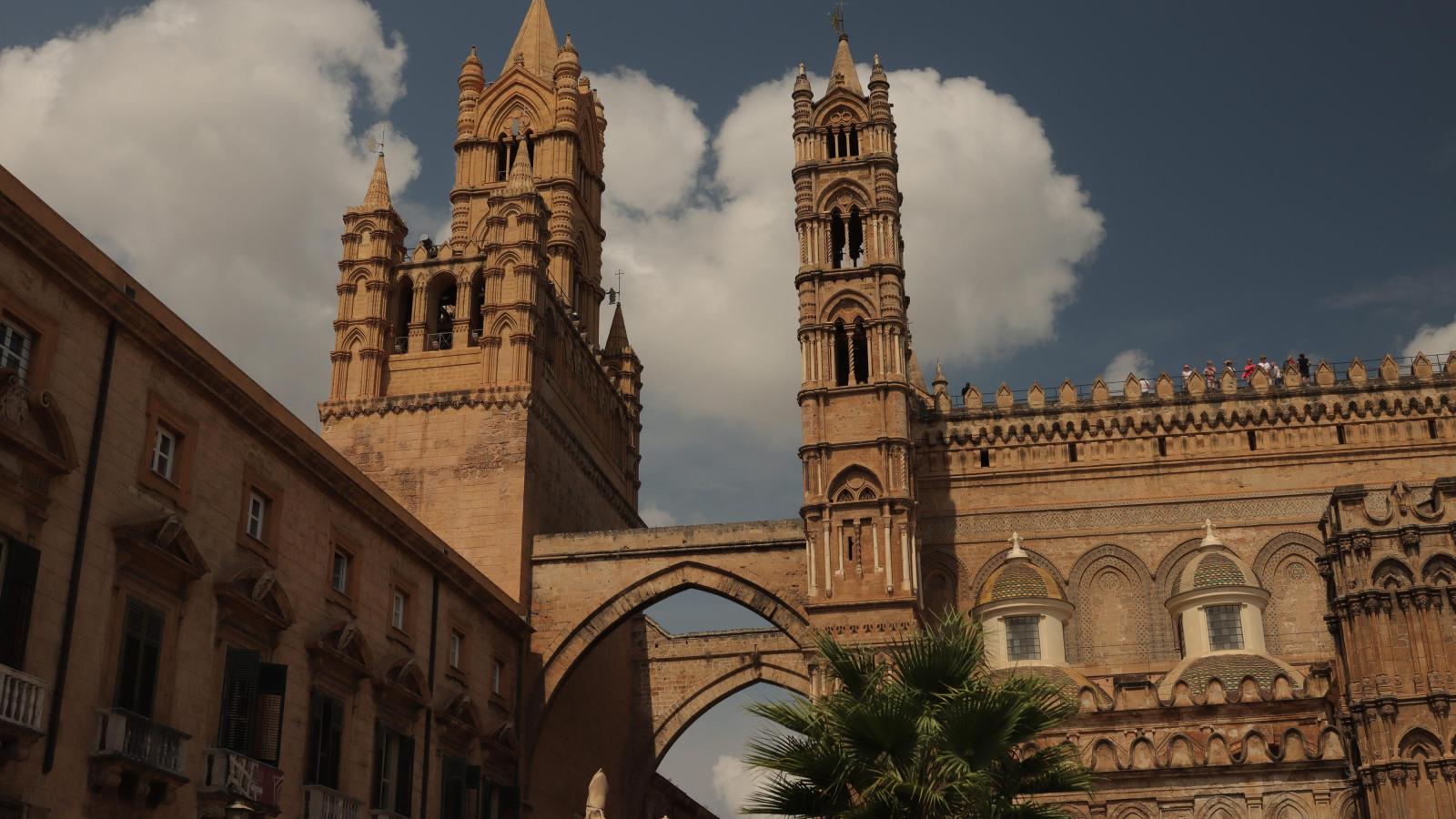 Vor einem blauen, leicht bew?lkten Himmeln ist die gro?e, alte Kathedrale von Palermo zu sehen. Sie ist aus hellem Stein. Zwei Trme strecken in die H?he und sind mit einem Torbogen verbunden.