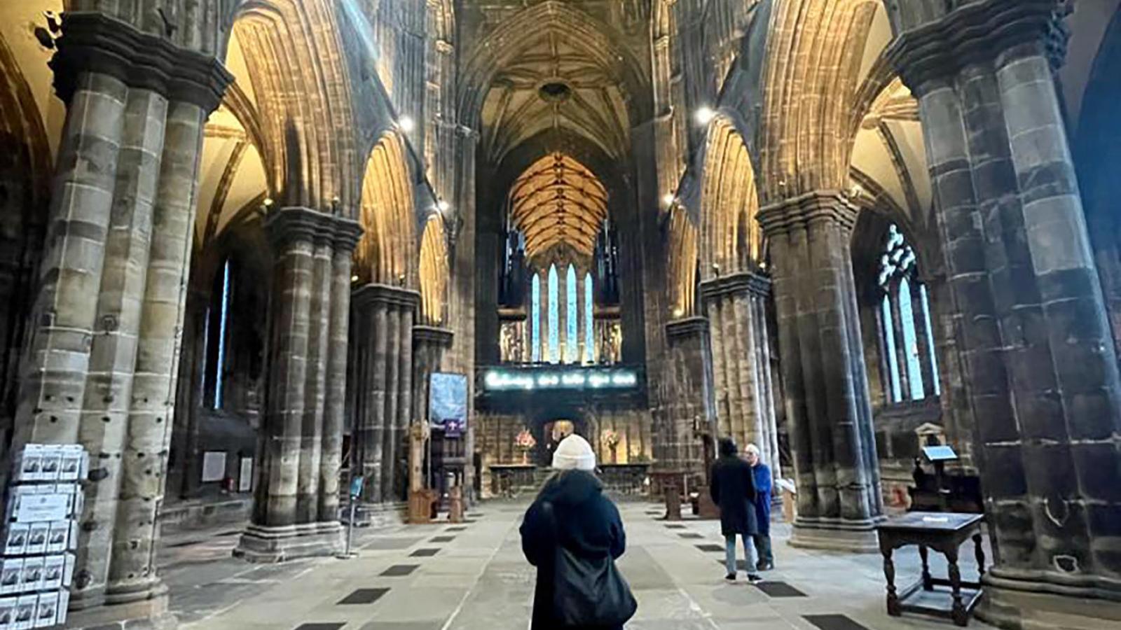 Ein Blick von innen in die Glasgower Kathedrale St. Mungo's Catherdral. Eine Person steht mittig mit dem Rcken zur Kamera. Der Raum ist Dunkel aber die Torbogen sind von unten beleuchtet.